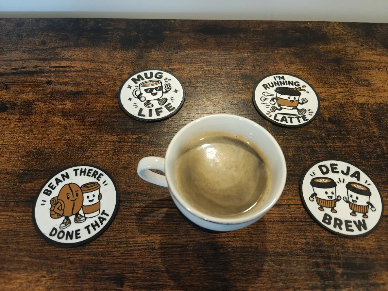 A real cup of coffee surrounded by four finished Quality3Ds coffee pun coasters on a wooden table.
