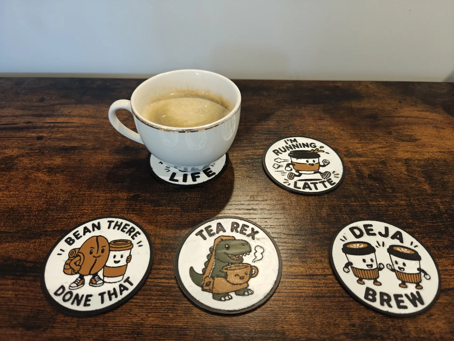 A real cup resting on a Quality3Ds coaster beside four finished coffee pun coaster designs.