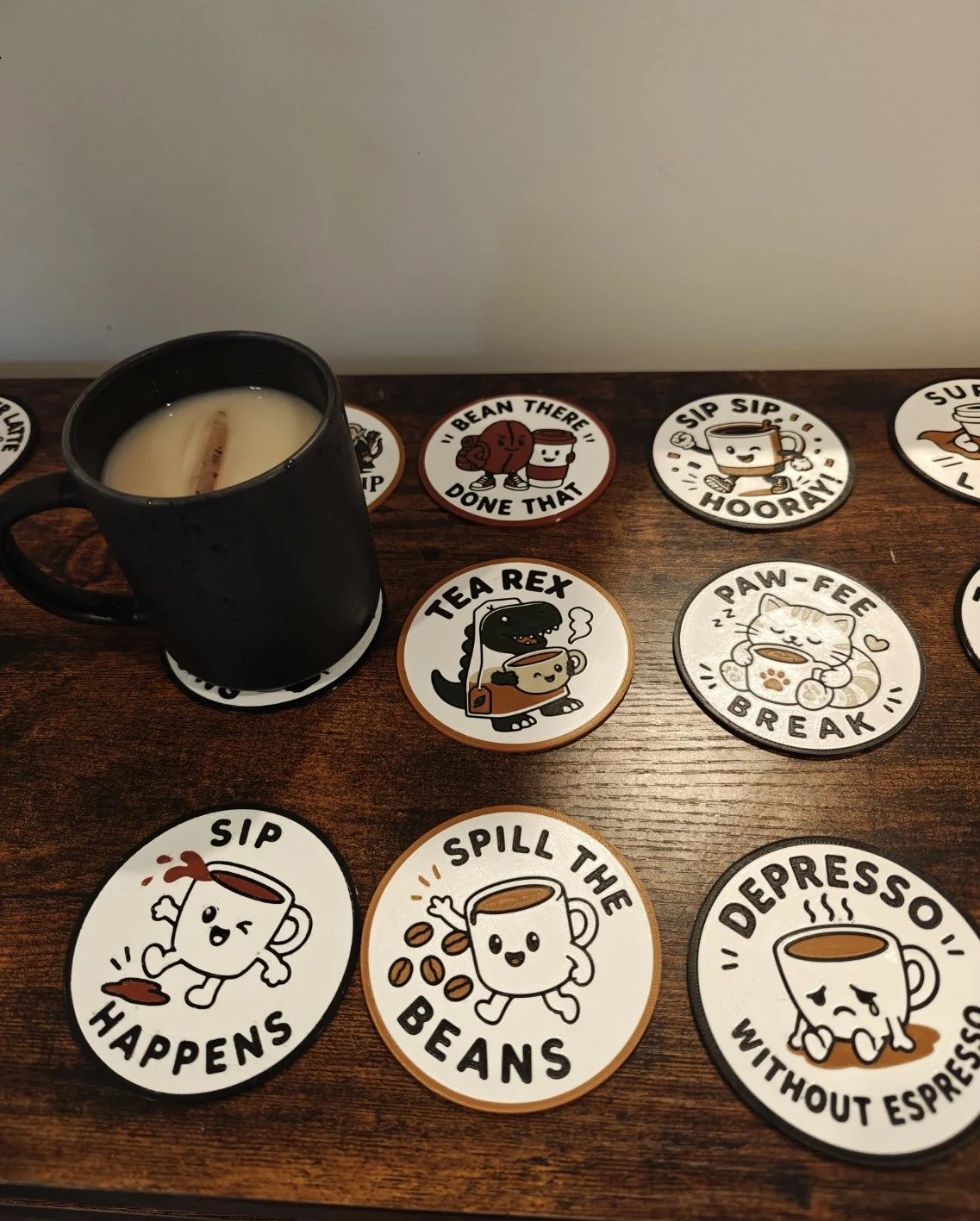 A black cup of tea surrounded by finished Quality3Ds coffee and tea pun coasters on a wooden table.
